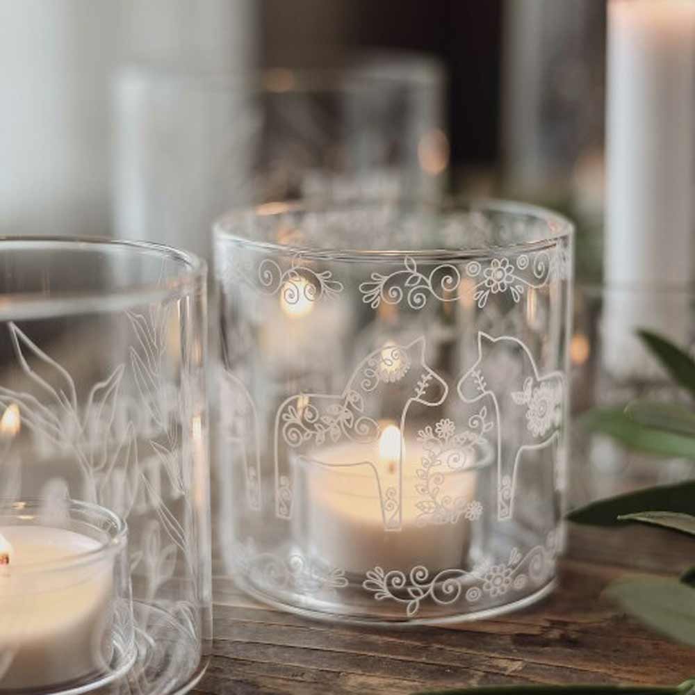 Majas Cottage - Teelichthalter Dala Pferde Glas Windlichter mit weißen Blumen- und Pferdemotiven enthalten brennende Teelichter; Im Hintergrund ist etwas Grün zu sehen.