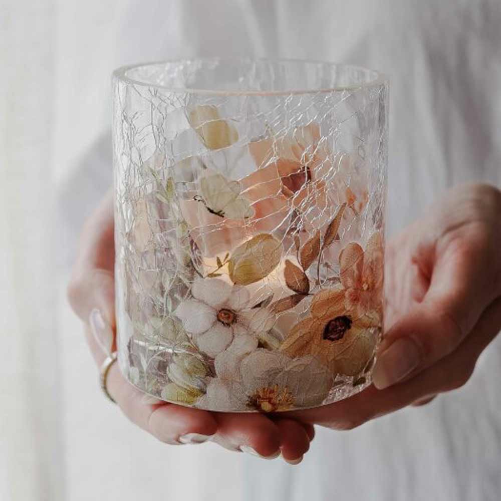Eine Person hält das Majas Cottage – Windlicht Blossom & Butterflies, einen Kerzenhalter aus klarem Glas, der mit gepressten Blumen, Schmetterlingen, zarten Rissen und einer brennenden Kerze darin geschmückt ist.
