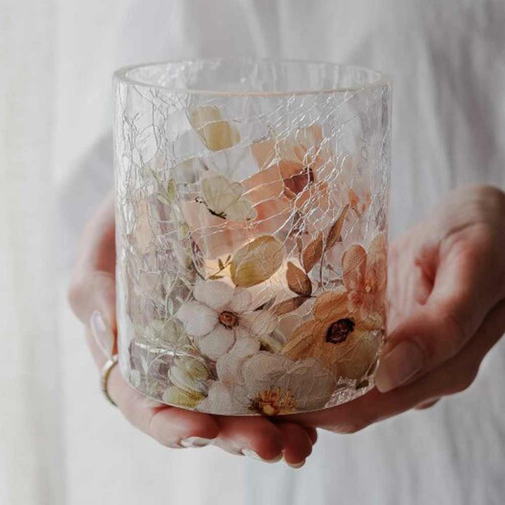 Eine Person hält das Majas Cottage – Windlicht Blossom & Butterflies, einen Kerzenhalter aus klarem Glas, der mit gepressten Blumen, Schmetterlingen, zarten Rissen und einer brennenden Kerze darin geschmückt ist.