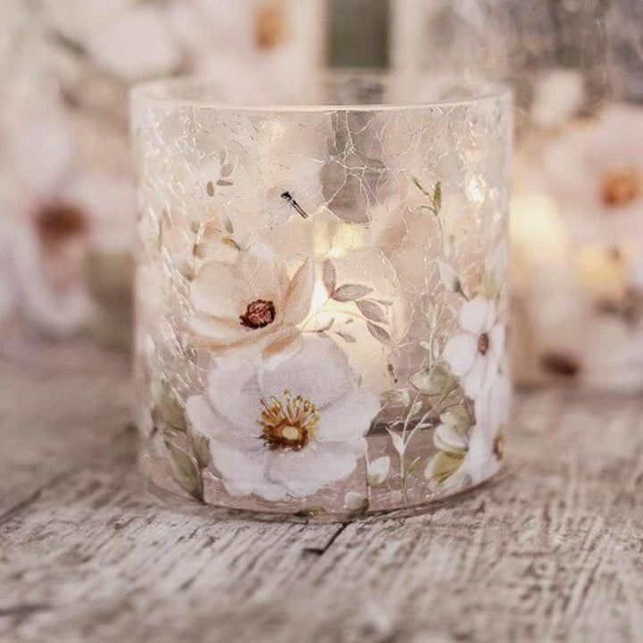Das Majas Cottage Windlicht Blossom & Butterflies, ein Kerzenhalter aus Craquelé-Glas mit weißen Blumenmustern, hält eine brennende Kerze und ruht auf einer rustikalen Holzoberfläche.