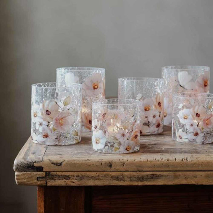Sechs Glaskerzenhalter „Windlicht Blossom & Butterflies“ von Majas Cottage mit eingelegten rosa und weißen Blumen sind auf einem rustikalen Holztisch vor einem neutralen Hintergrund angeordnet.