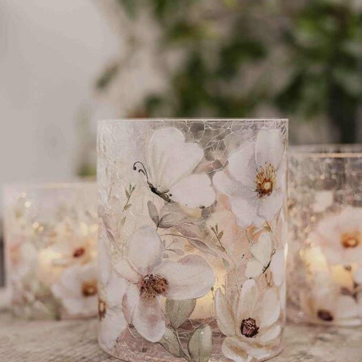 Das Majas Cottage – Windlicht Blossom & Butterflies sind drei Kerzenhalter aus Glas mit weißen Blumen- und Schmetterlingsmotiven, die jeweils von einer kleinen Kerze beleuchtet werden und auf einer hellen Oberfläche mit verschwommenem Grün im Hintergrund stehen.