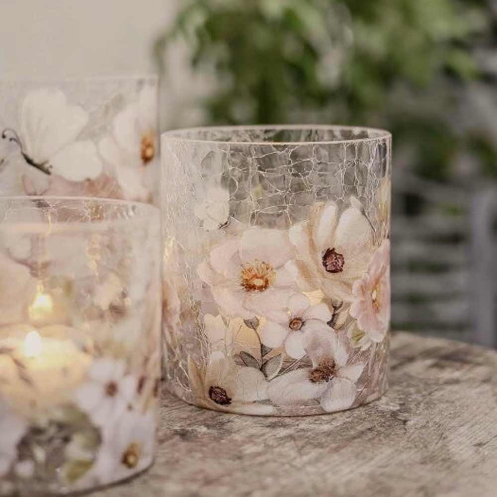 Drei Glaskerzenhalter „Windlicht Blossom & Butterflies“ von Majas Cottage mit Blumenmuster stehen auf einer Holzoberfläche und halten jeweils eine brennende Kerze, während im Hintergrund sanft verschwimmendes Grün zu sehen ist.