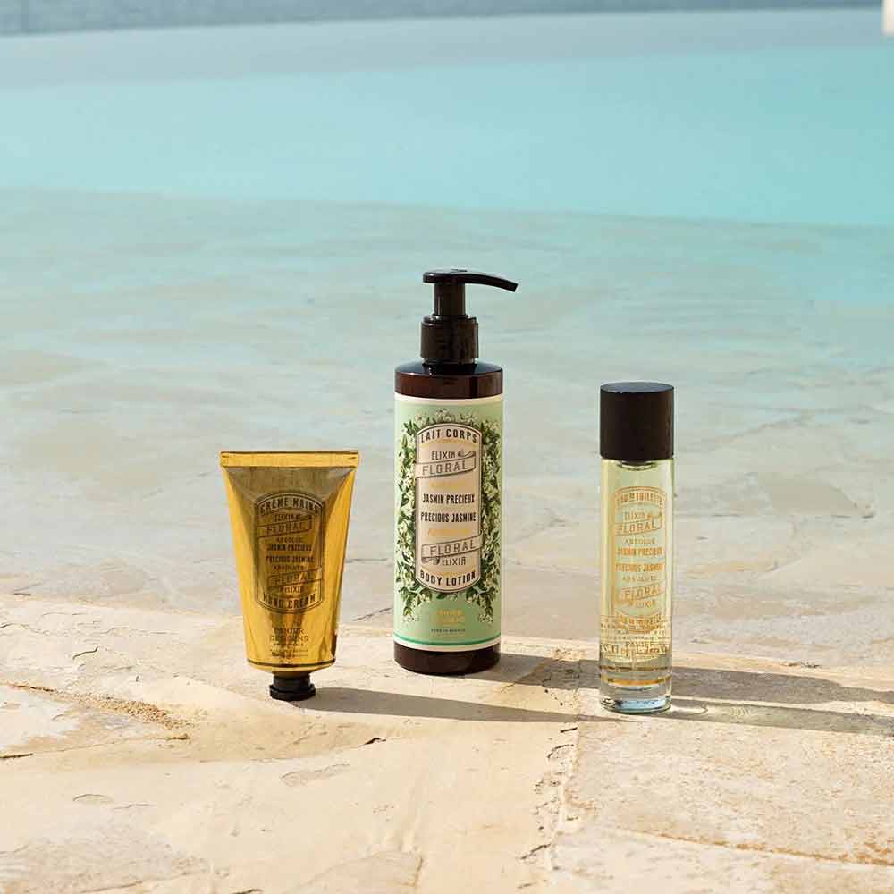 Drei Hautpflegeprodukte mit Etiketten im Vintage-Stil sind auf einer beigen Steinoberfläche in der Nähe eines Pools angeordnet. Dazu gehören eine goldene Tube, Panier des Sens – Bodylotion Jasmin 250 ml, und eine kleine Sprühflasche aus Glas.