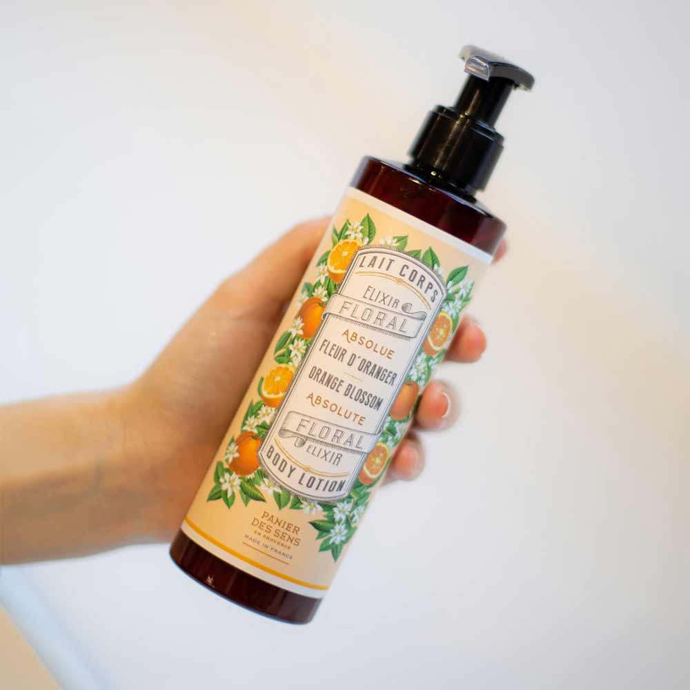 Eine Hand hält eine Flasche Bodylotion mit Pumpspender. Auf dem Etikett steht „Panier des Sens – Bodylotion Orangenblüte 250ml“. Die Flasche ist mit Bildern von Orangen und grünen Blättern verziert.