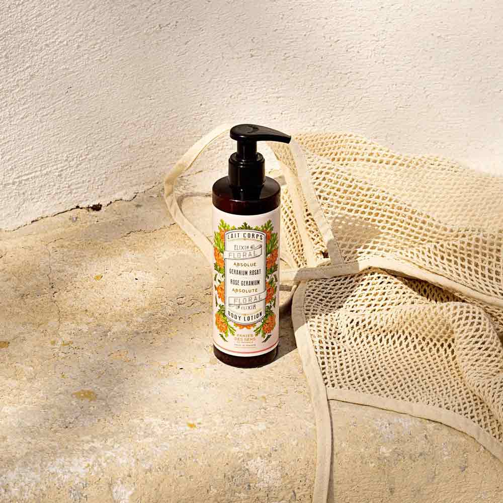 Eine braune Pumpflasche Panier des Sens - Bodylotion Rosengeranie 250ml steht aufrecht auf einer Steinfläche neben einem beigen Netzbeutel. Im Hintergrund ist eine weiße Wand im Sonnenlicht zu sehen.