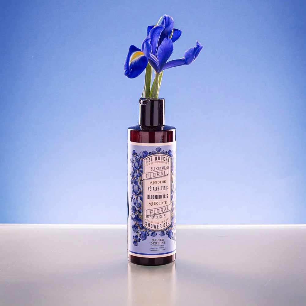 In einer braunen Kosmetikflasche mit der Aufschrift „Panier des Sens – Duschgel Irisblütenblätter 250 ml“ ist vor blauem Hintergrund eine blaue Irisblüte platziert.
