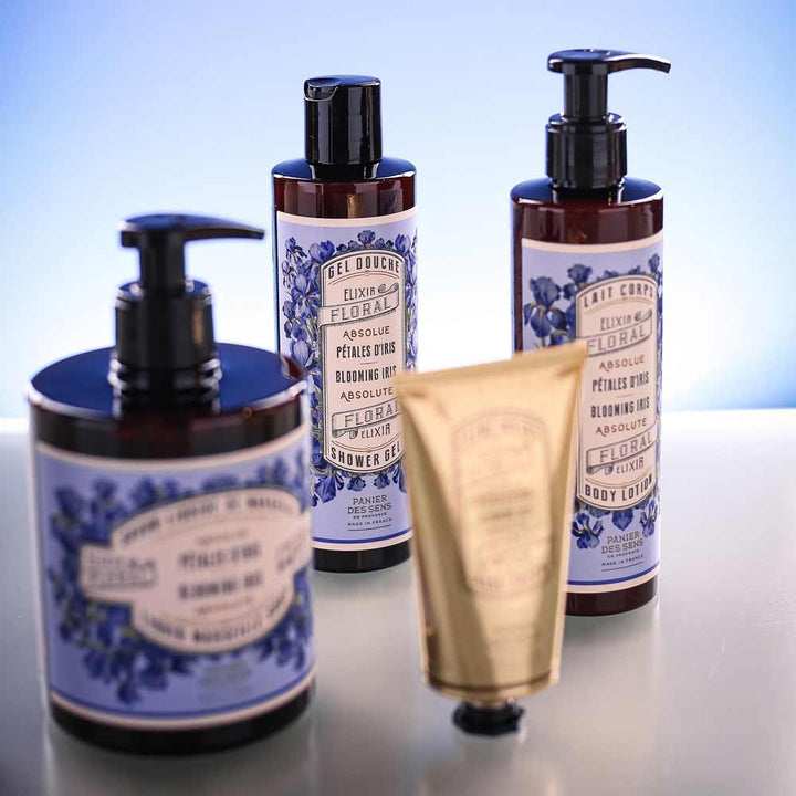 Ausgestellt werden vier Hautpflegeprodukte, darunter drei Pumpflaschen und ein Panier des Sens - Duschgel Irisblütenblätter 250 ml. Alle haben Etiketten mit Blumenmotiven und violetten Schattierungen und goldenen Akzenten. Der Hintergrund ist sanft blau beleuchtet.