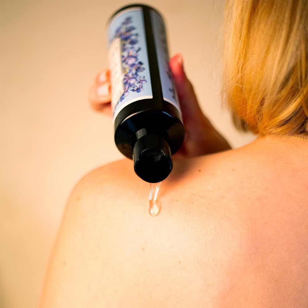 Eine Person mit blonden Haaren gießt Panier des Sens – Duschgel Irisblütenblätter 250ml auf ihre nackte Schulter.