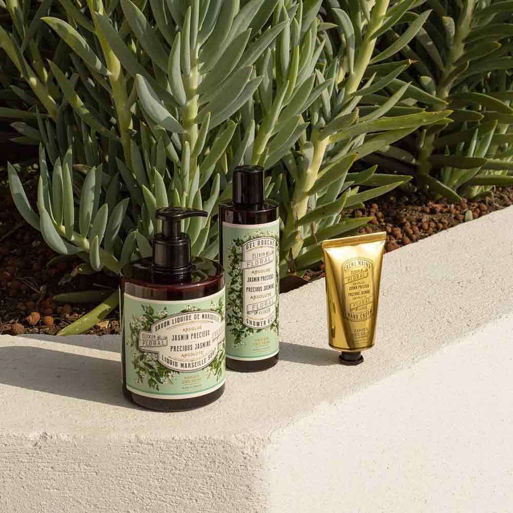 Drei Behälter mit 250 ml „Panier des Sens – Duschgel Jasmin“, darunter zwei Pumpflaschen und eine kleine goldene Tube, werden auf einem Betonsims mit grünem Laub im Hintergrund präsentiert.