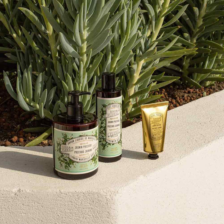 Drei Behälter mit 250 ml „Panier des Sens – Duschgel Jasmin“, darunter zwei Pumpflaschen und eine kleine goldene Tube, werden auf einem Betonsims mit grünem Laub im Hintergrund präsentiert.