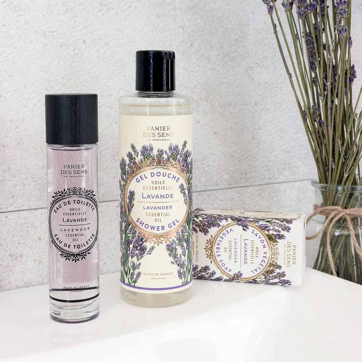 Ein Set Panier des Sens – Duschgel Lavendel 250 ml auf einer weißen Fläche, bestehend aus einer Parfümflasche, Duschgel und Seife, mit Lavendelzweigen im Hintergrund.