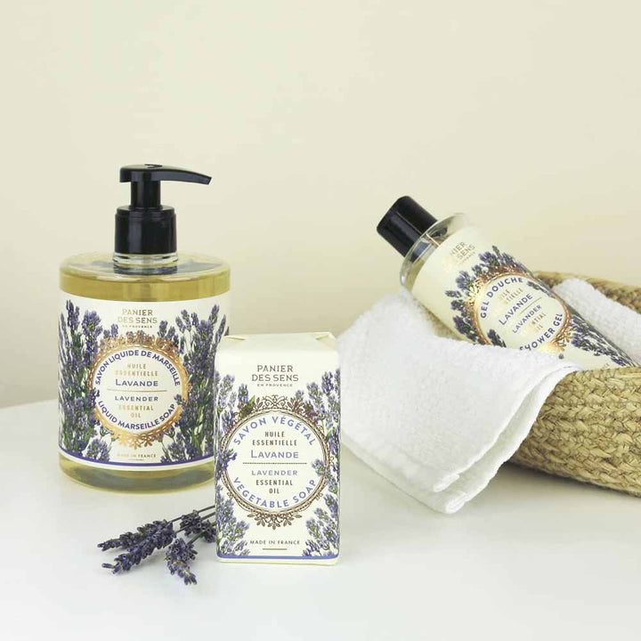 Eine Sammlung von Badeprodukten mit Lavendelduft, darunter Panier des Sens – Duschgel Lavendel 250 ml, Handcreme und ein Stück Seife, sind auf einer weißen Fläche neben einem Weidenkorb mit Handtüchern angeordnet.