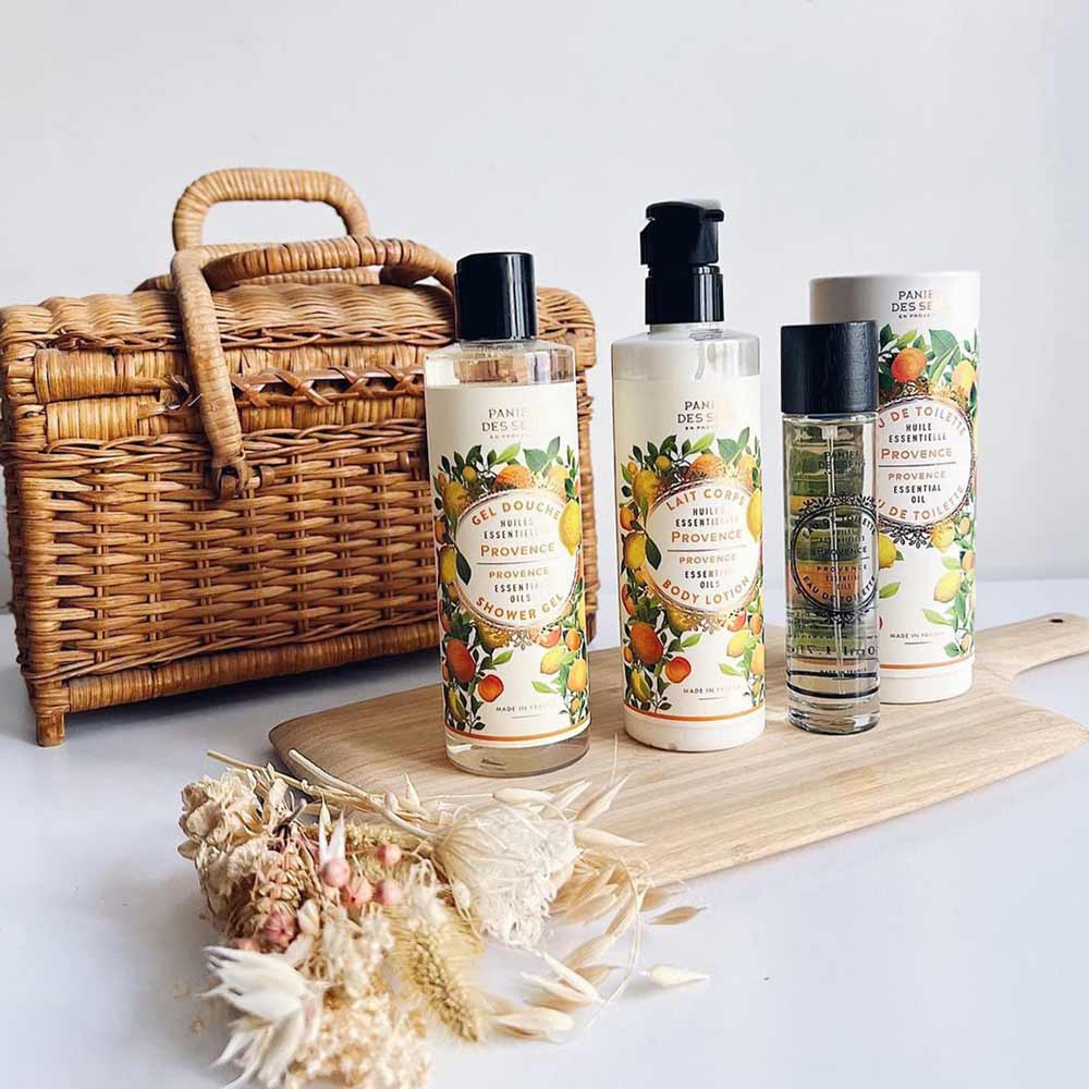 Ein Weidenkorb, ein Holzbrett und getrocknete Blumen werden neben vier Bade- und Körperprodukten mit Zitrusduft ausgestellt, darunter ein Panier des Sens – Duschgel Provence 250 ml, Körperlotion, Handcreme und Parfüm.