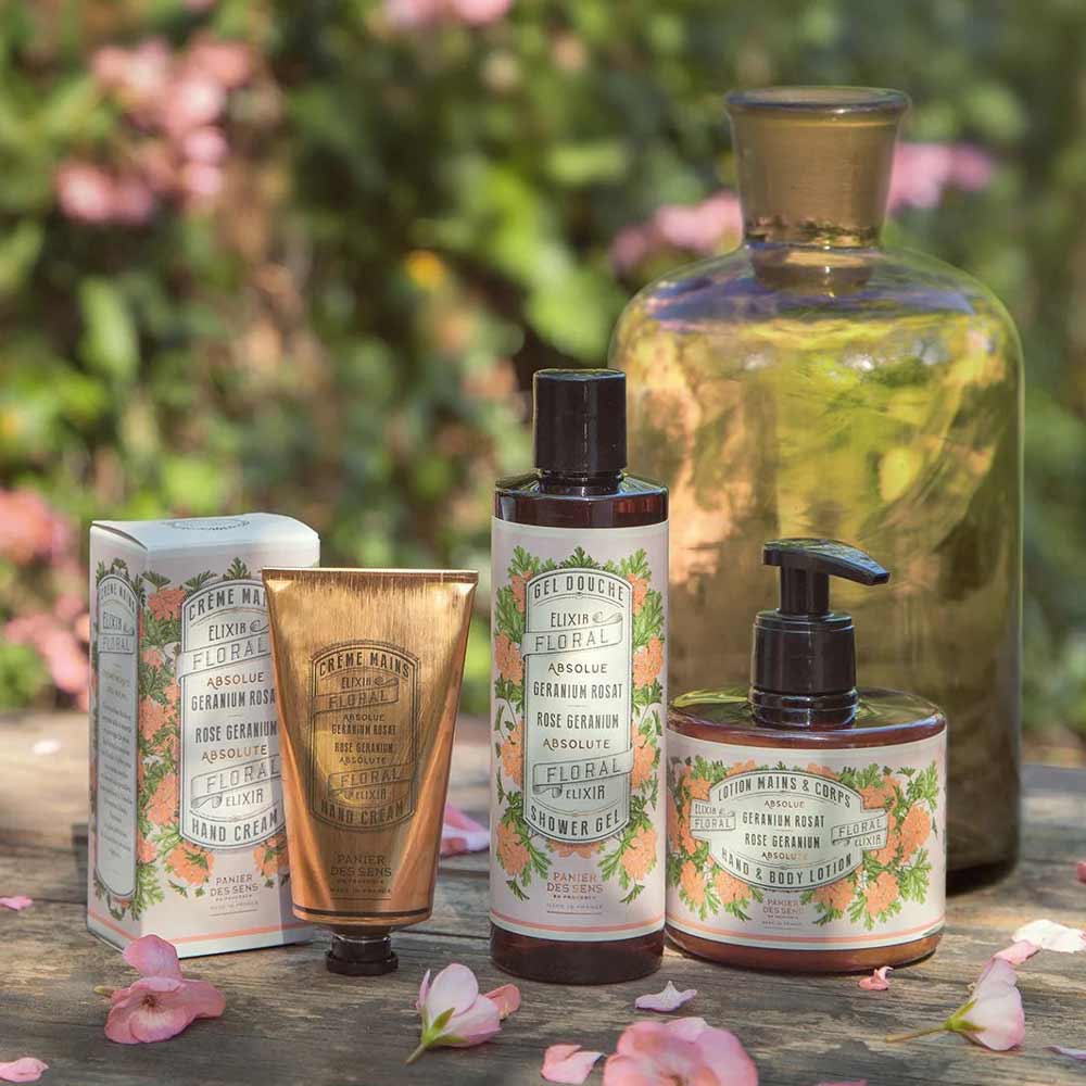 Eine Kollektion von Hautpflegeprodukten, darunter eine Handcreme, Panier des Sens - Duschgel Rosengeranie 250 ml, Körperlotion und Seife, im Freien arrangiert mit rosa Blumen im Hintergrund. Jedes Produkt hat eine Verpackung mit Blumenmotiv.