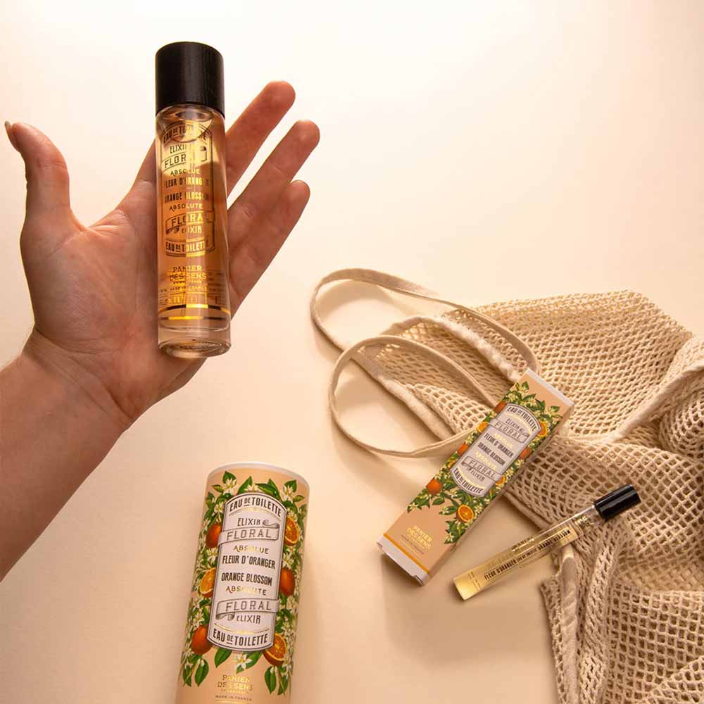 Eine Hand hält einen Panier des Sens – Eau de Toilette Roll-on Orangenblüte 10ml. Daneben liegen auf einer beigen Fläche ein Netzbeutel, zwei Parfümschachteln und ein weiteres Parfümfläschchen.