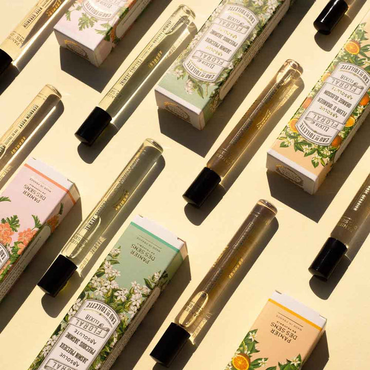 Verschiedene Parfümflakons und -schachteln sind flach auf einer hellen Oberfläche angeordnet. Die Schachteln sind mit Blumenmotiven und Textbeschreibungen der Düfte versehen, darunter Panier des Sens – Eau de Toilette Roll-on Orangenblüte 10 ml.