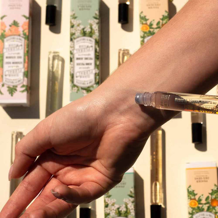 Eine Person trägt einen Panier des Sens – Eau de Toilette Roll-on Orangenblüte 10ml auf ihr Handgelenk auf. Im Hintergrund sind verschiedene Flakons und Schachteln mit Düften zu sehen.