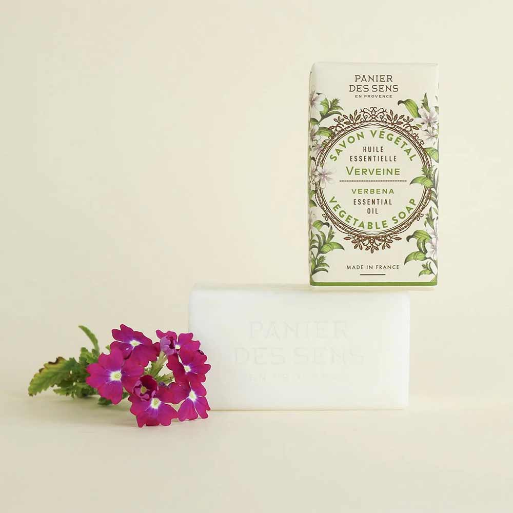 Ein Stück Panier des Sens - Feste Pflanzenseife Eisenkraut (Verbene) 150 g Seife und ihre Verpackung von Panier des Sens mit Eisenkraut, dargestellt mit einer rosa Blume auf beigem Hintergrund.