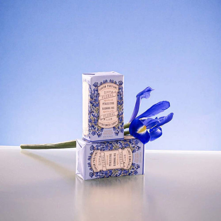 Zwei Riegel à 150 g „Panier des Sens – Feste Seife mit Duft Irisblütenblätter“ sind auf einem hellblauen Hintergrund gestapelt, begleitet von einer einzelnen blauen Irisblüte.