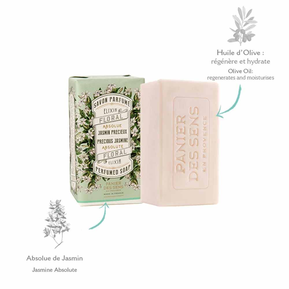 Ein Stück Jasminseife von Panier des Sens neben der Verpackung mit der Aufschrift „Elixir Floral Absolue de Jasmin“ und einer Liste der wohltuenden Wirkungen von Olivenöl vor einem schlichten Hintergrund.