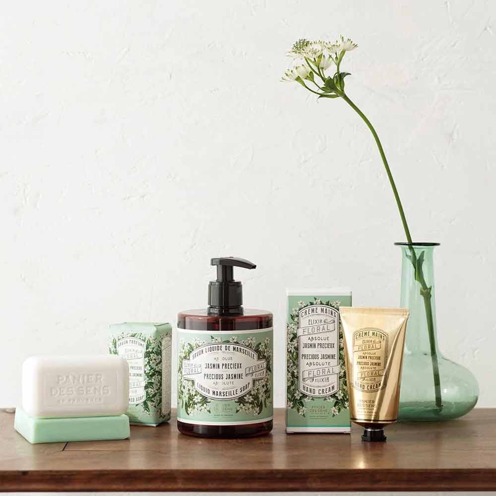 Eine Kollektion französischer Jasminseifenprodukte von Panier des Sens, darunter Seife, Flüssigseife und Creme, präsentiert neben einer grünen Vase mit einer einzelnen Blume.