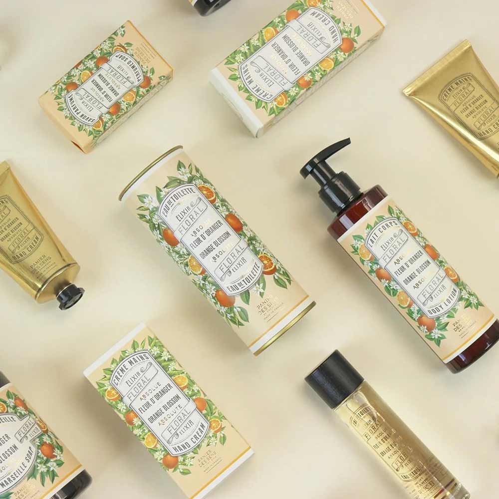 Verschiedene Körperpflegeprodukte, darunter Lotionen, Cremes und die Panier des Sens - Feste Seife mit Duft Orangenblüte 150g, sind auf einem cremefarbenen Hintergrund angeordnet. Alle Produkte verfügen über eine Verpackung im Vintage-Stil mit Blumen- und Fruchtillustrationen.