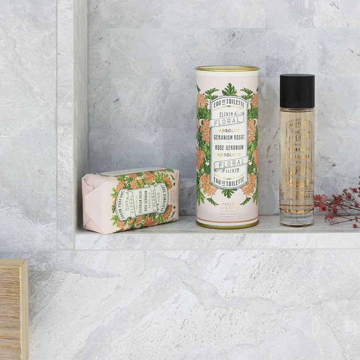 Ein Panier des Sens – Feste Seife mit Duft Rosengeranie 150 g, ein zylindrischer Behälter und eine kleine Sprühflasche sind auf einem Badezimmerregal vor einem grauen Marmorhintergrund angeordnet.