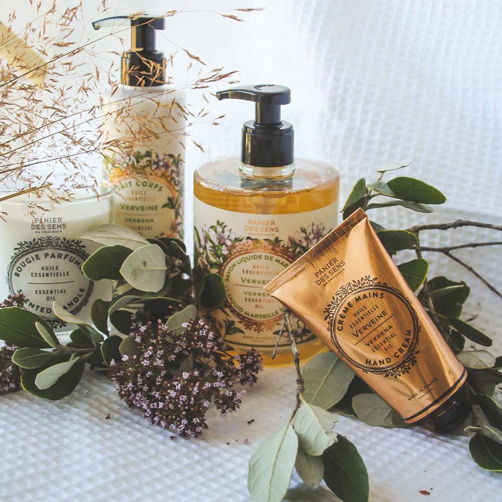 Ein Arrangement von Panier des Sens - Handcreme Eisenkraut (Verbene) mit einem natürlichen Hintergrund aus Blättern und Blüten.