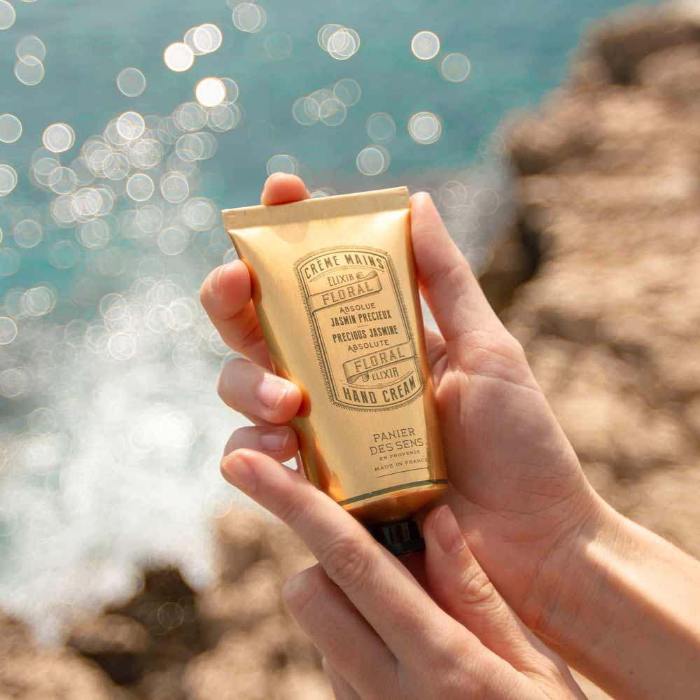 Eine Person hält Panier des Sens - Handcreme Jasmin vor glitzerndem Wasser im Hintergrund.