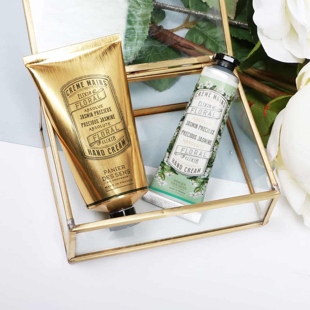 Zwei Tuben Panier des Sens – Handcreme Jasmin, präsentiert in einer eleganten Vitrine aus Glas und Gold, mit weißen Blumen im Hintergrund.