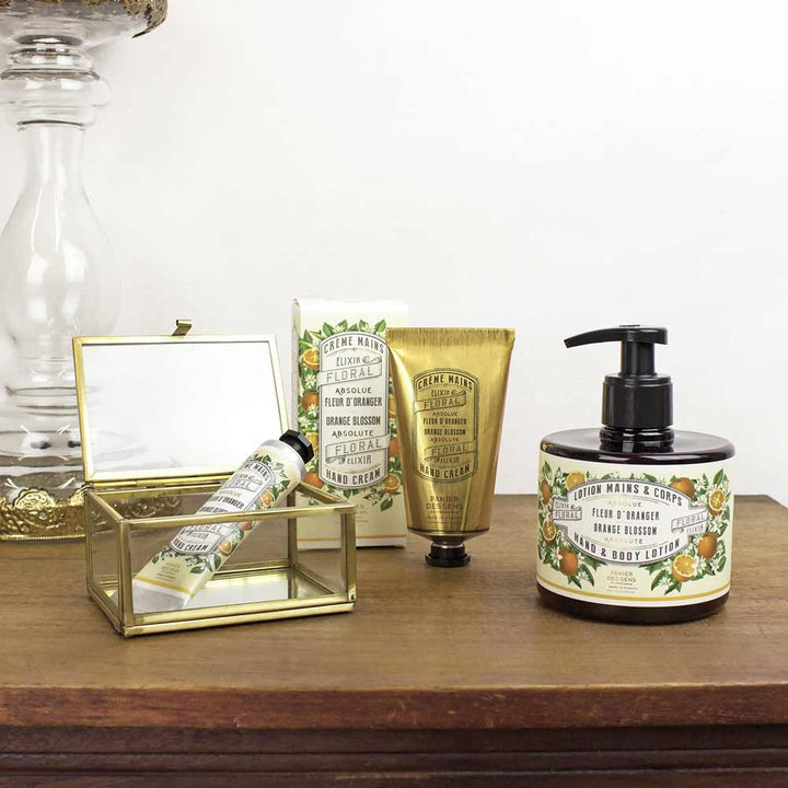 Eine Reihe von Panier des Sens - Handcreme Orangenblüte, arrangiert auf einer Holzoberfläche neben einer goldgerahmten Glasbox.