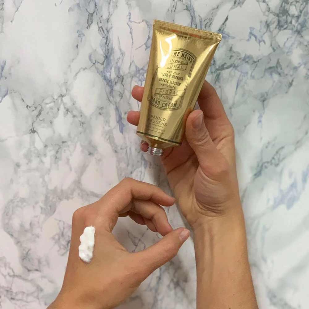 Eine Person hält eine Tube Panier des Sens - Handcreme Orangenblüte mit einem Tupfer des Produkts auf ihrem Finger vor einem Marmorhintergrund.