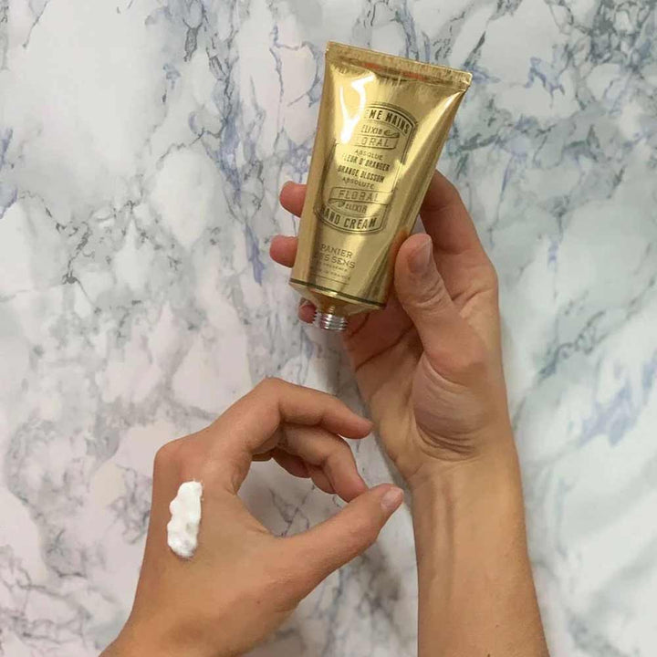 Eine Person hält eine Tube Panier des Sens - Handcreme Orangenblüte mit einem Tupfer des Produkts auf ihrem Finger vor einem Marmorhintergrund.