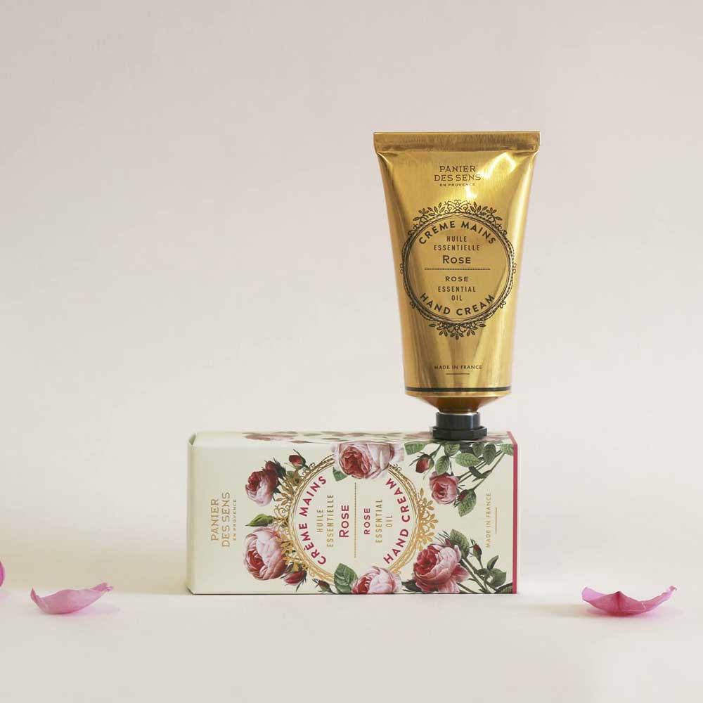 Eine Tube Panier des Sens - Handcreme Rose mit Verpackung auf beigem Hintergrund, begleitet von verstreuten Rosenblättern.