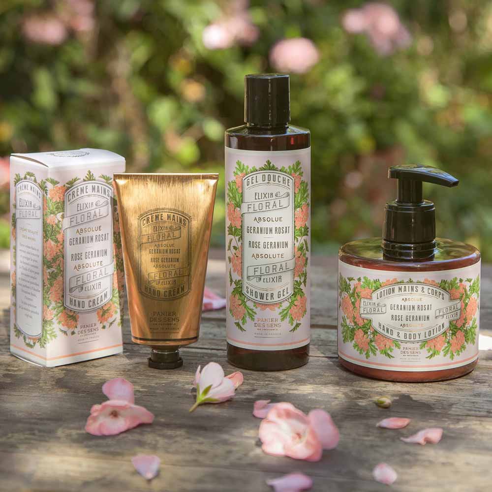 Sortiment von Panier des Sens - Handcreme Rosengeranie auf einer von Blütenblättern umgebenen Holzoberfläche.