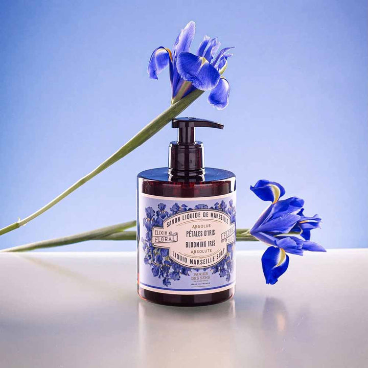 Eine Flasche Panier des Sens – Marseiller Flüssige Handseife Irisblütenblätter 500 ml mit schwarzem Pumpverschluss, verziert mit blauen Blumengrafiken, platziert auf einer Oberfläche mit blauen Schwertlilien im Hintergrund vor einem hellblauen Hintergrund.