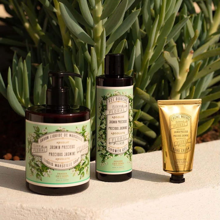 Drei Hautpflegeprodukte, darunter Panier des Sens – Marseiller Flüssige Handseife Jasmin 500 ml, ein Duschgel und eine Handcreme, werden vor einer grünen Pflanze ausgestellt. Die Produkte haben grüne Etiketten mit Blumenmustern und eine verzierte goldene Tube.