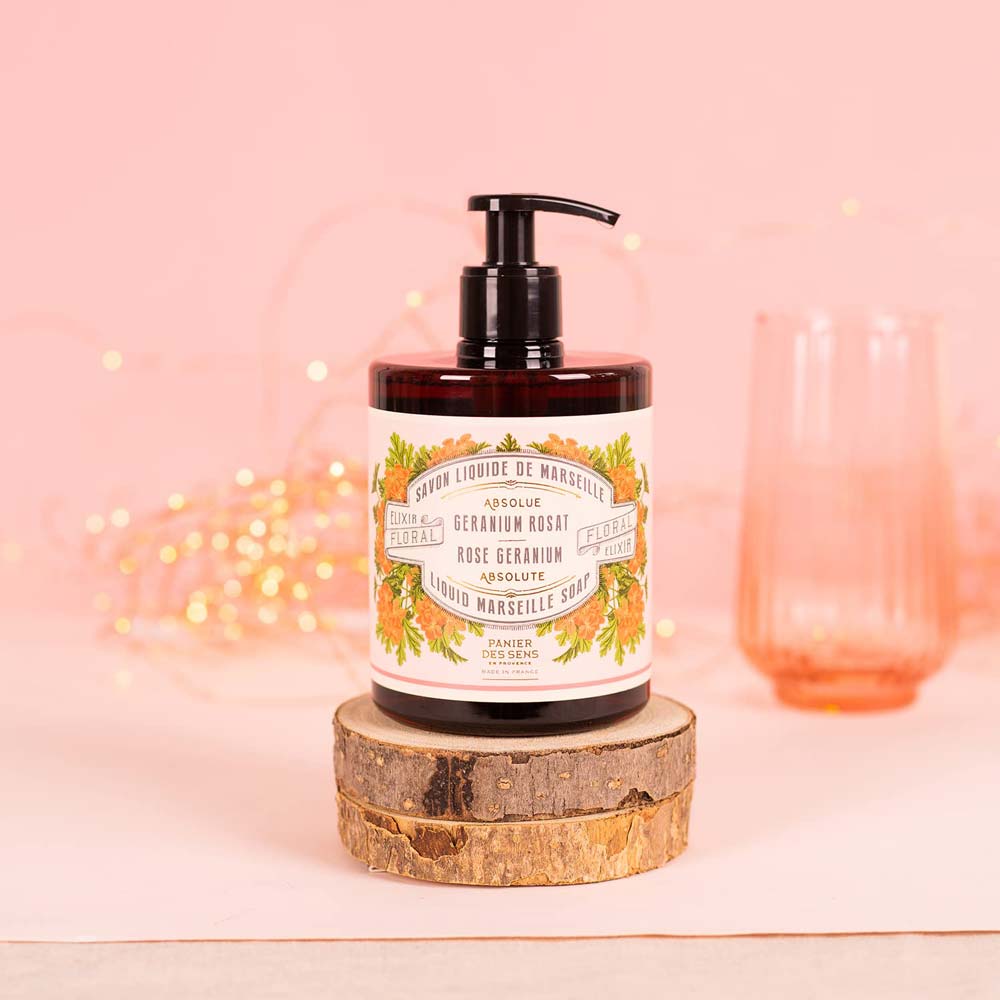 Eine Flasche Panier des Sens – Marseiller Flüssige Handseife Rosengeranie 500 ml mit Blumenetikett und Pumpspender steht auf einem Holzständer. Zu sehen ist ein rosa-orangefarbener Hintergrund sowie ein durchscheinendes Glas.