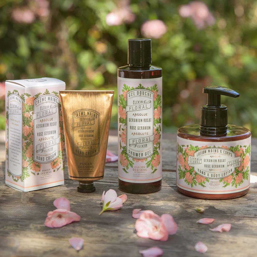 Vier Hautpflegeprodukte mit Blumenmotiven, darunter Panier des Sens – Marseiller Flüssige Handseife Rosengeranie 500 ml, Duschgel und Körperlotion, werden auf einer Holzoberfläche mit rosa Blütenblättern drumherum präsentiert.