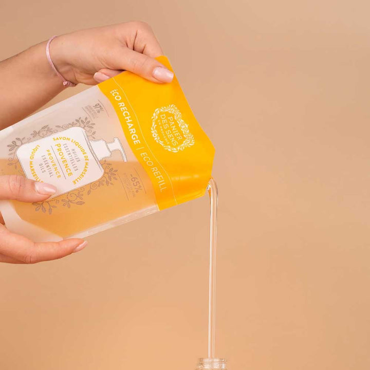 Eine Person gießt Panier des Sens - Marseiller Flüssigseife Nachfüllbeutel Provence 500 ml aus einer bernsteinfarbenen Flasche in einen kleinen Behälter vor beigem Hintergrund.