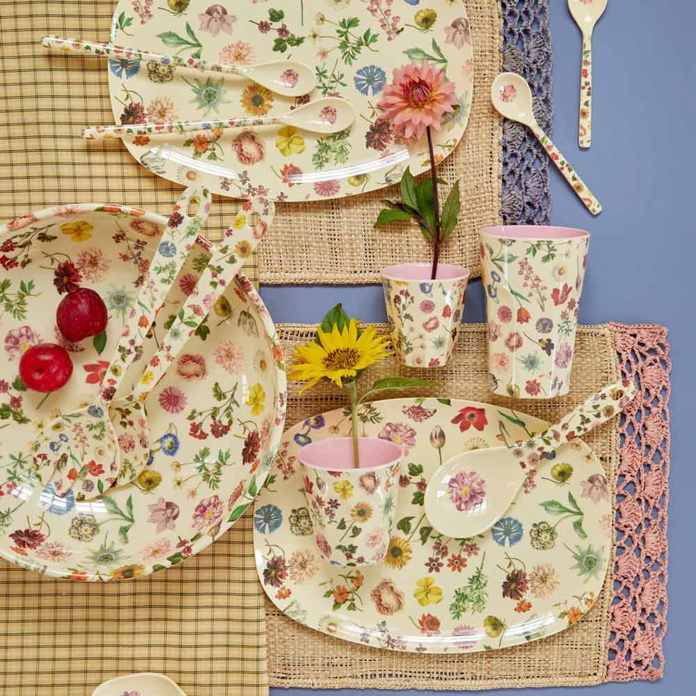 Eine Tischdekoration mit Tellern, Schüsseln, Löffeln mit Blumenmuster, Reis-Melaminbecher Floras Dream Medium und zwei Blumen in Bechern. Zwei Radieschen befinden sich in einer Schüssel, darunter karierte und gewebte Tischsets.