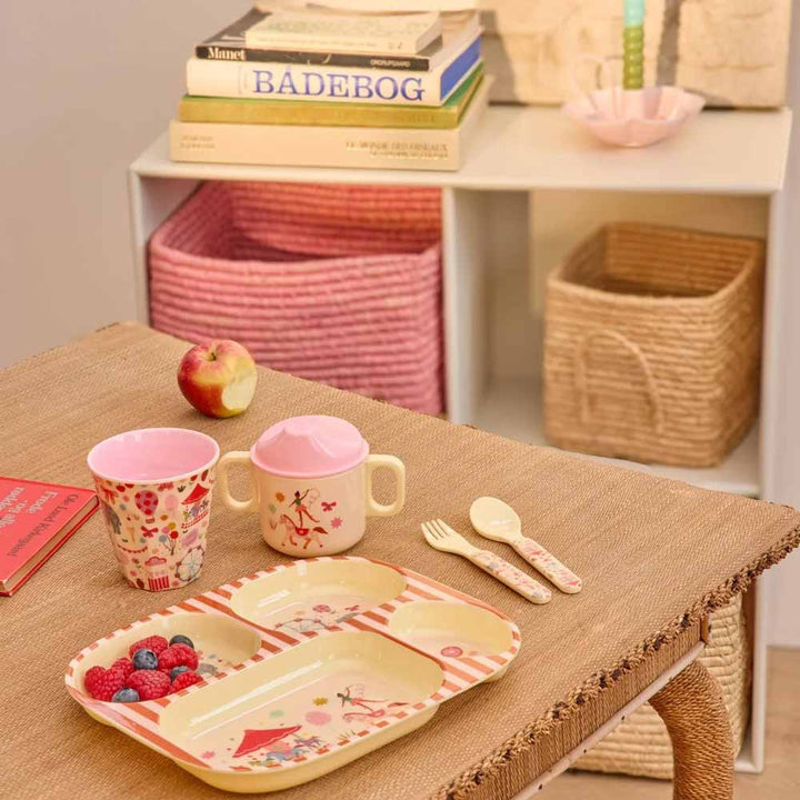 Auf einem geflochtenen Tisch steht ein Kinderteller mit Obst, Besteck und einem Rice Melaminbecher Fun Fair Medium. Im Hintergrund stehen Körbe und Bücher in den Regalen, und auf dem Tisch liegt ein Apfel.