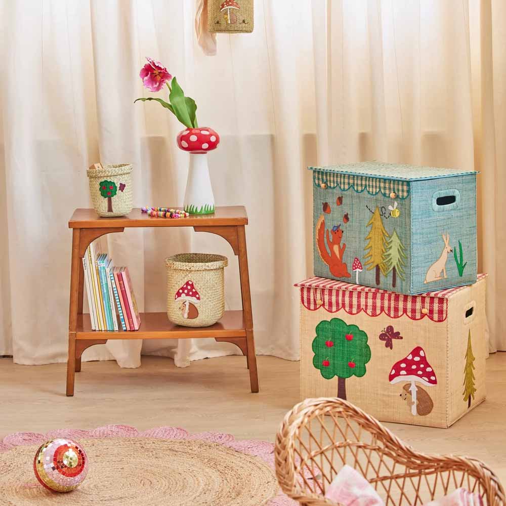 Ein Kinderzimmer mit dem Rice - Raffia Aufbewahrungskorb Happy Forest Faltbar mit Tier- und Pilzmotiven, einem kleinen Holzregal mit Büchern, einer Kunstblume, einem Pilzornament und einem runden gewebten Teppich.