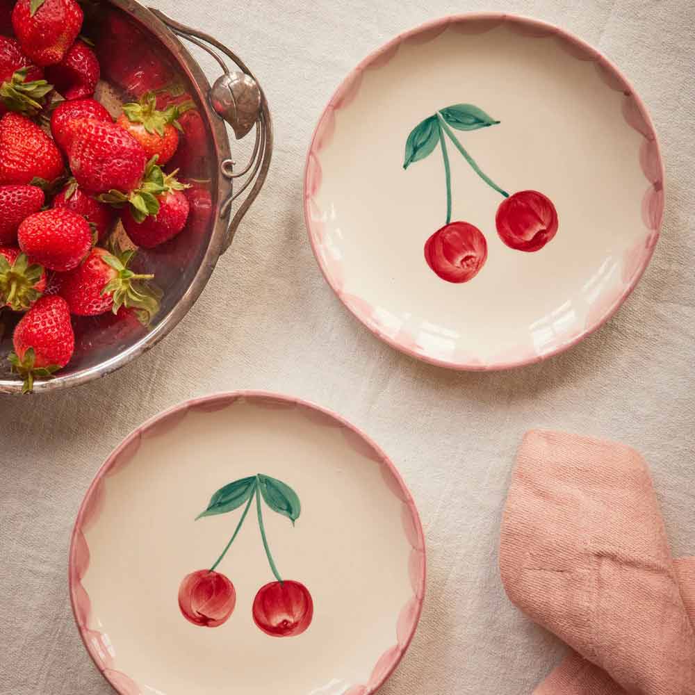 Zwei Keramikteller von Rice mit handgemalten Kirschen stehen neben einer Metallschale mit Erdbeeren und einer rosa Serviette auf einer weißen Tischdecke.
