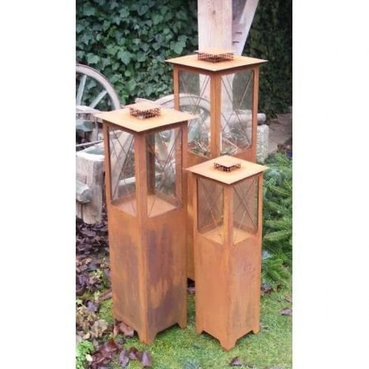 Die Rostikal Edelrost Metall Laterne Windlaterne Mega 105 cm steht im Freien mit Glasscheiben, umgeben von Gras, Blättern, einem Holzrad und Grünzeug.