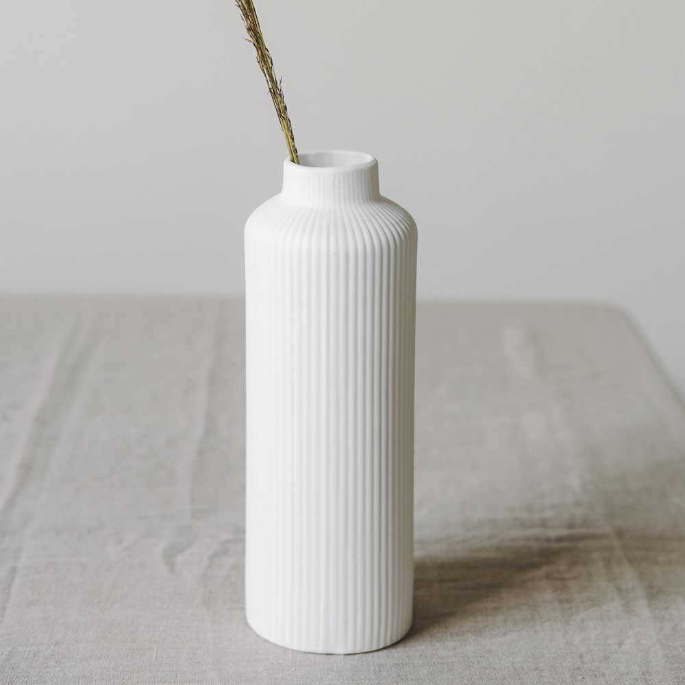 Die Ådala-Vase von Storefactory aus weißer Keramik mit geripptem Design steht elegant mit einem einzelnen getrockneten Zweig auf einer hellgrauen Oberfläche.