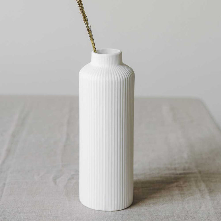 Die Ådala-Vase von Storefactory aus weißer Keramik mit geripptem Design steht elegant mit einem einzelnen getrockneten Zweig auf einer hellgrauen Oberfläche.