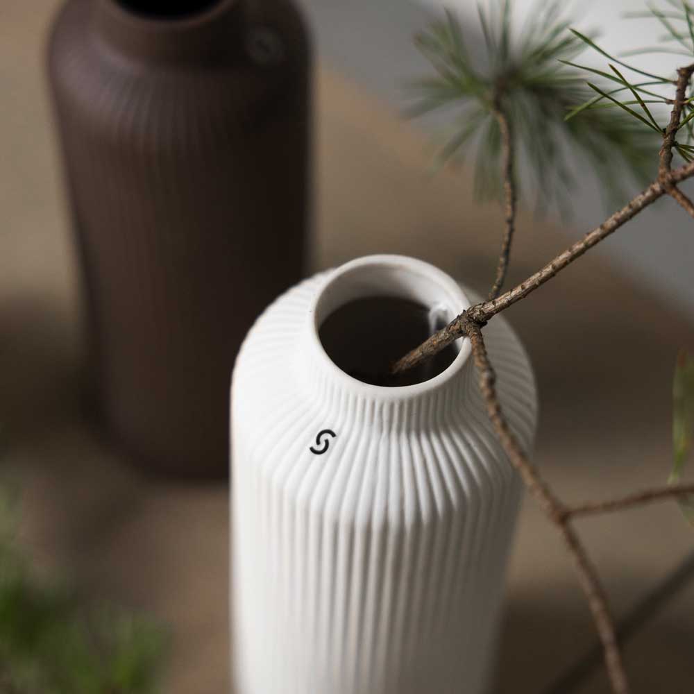 Zwei gerippte Keramikvasen von Storefactory – Ådala, eine weiße und eine braune, stehen auf einer Fläche. Ein Zweig mit grünen Nadeln ziert die weiße Vase.