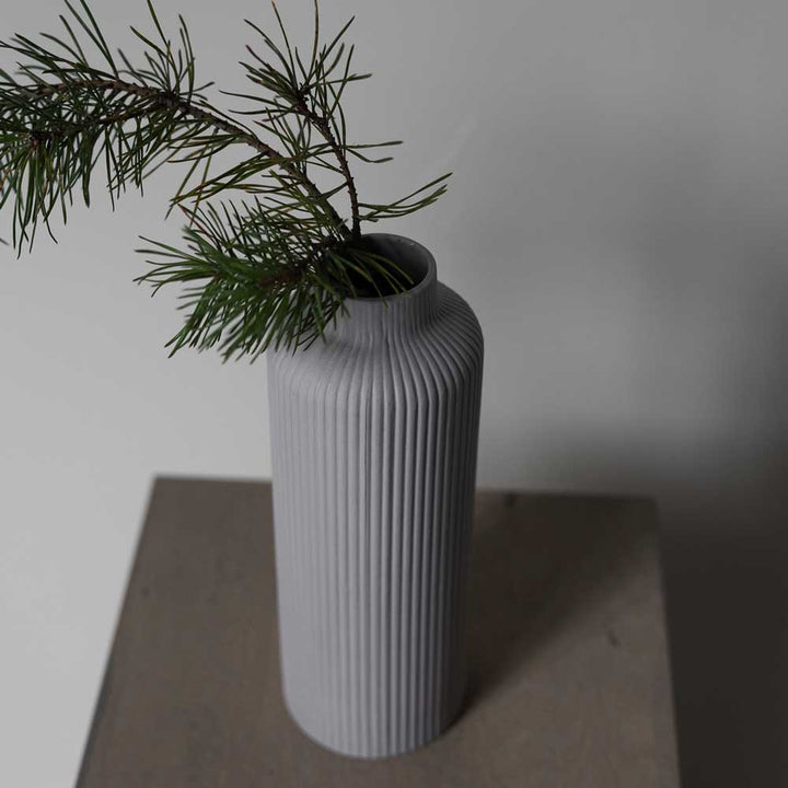 Die graue Keramikvase Ådala von Storefactory hält einen Zweig grüner Kiefernnadeln auf einer gedeckt braunen Oberfläche vor grauem Hintergrund.